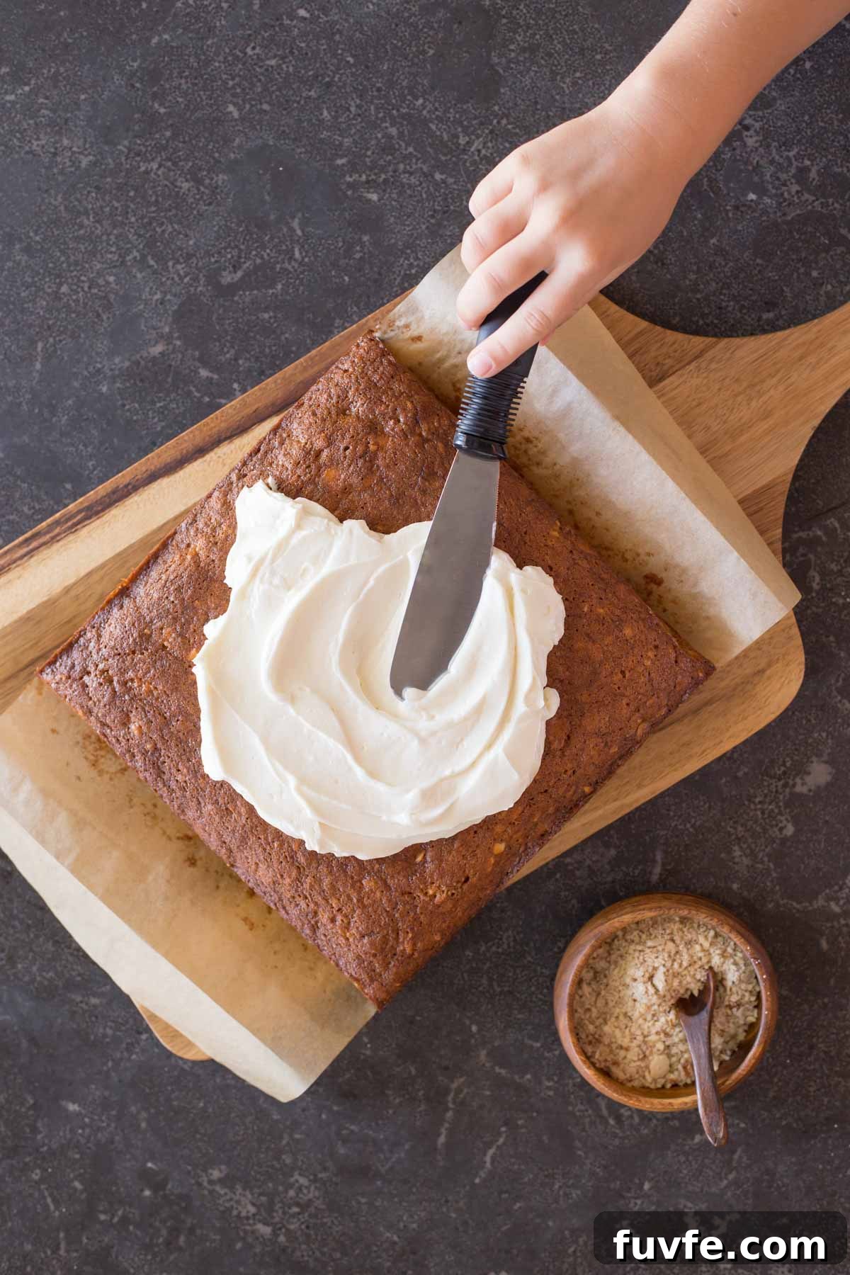 Banana Cake on parchment paper on top of a cutting board, with the Fluffy Cream Cheese Frosting being spread on top of the cake, and a small wood bowl of crushed almonds sitting next to the cutting board. 