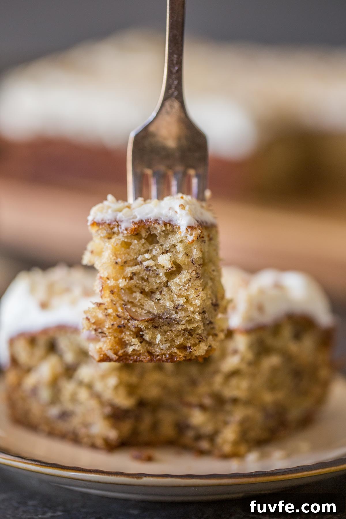 A fork with a bite of the Banana Cake with Fluffy Cream Cheese Frosting on it, with the rest of the piece of cake on a plate in the background.
