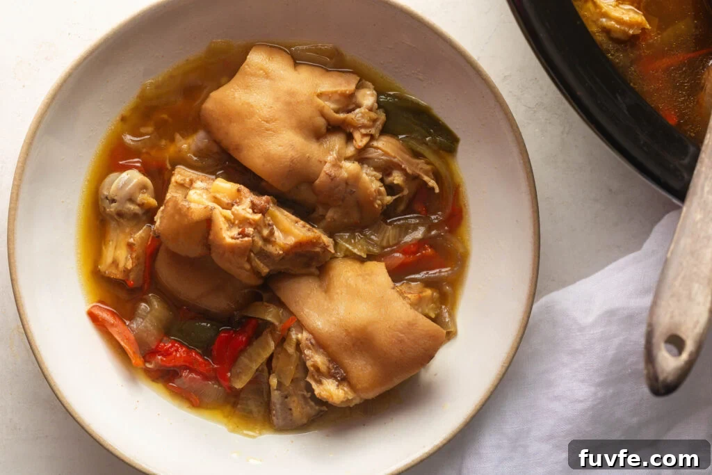 Bowl of cooked Pigs Feet with vegetables and braising liquid on white background with a white napkin next to it.