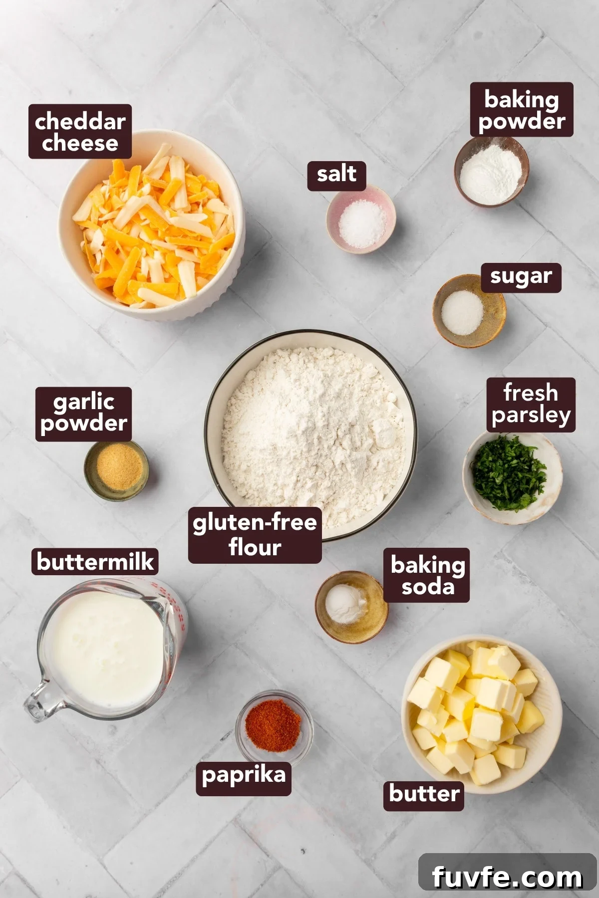 A flat lay of labeled ingredients for gluten-free cheddar biscuits, including cheddar cheese, salt, baking powder, sugar, fresh parsley, gluten-free flour, baking soda, butter, paprika, buttermilk, and garlic powder on a white surface.
