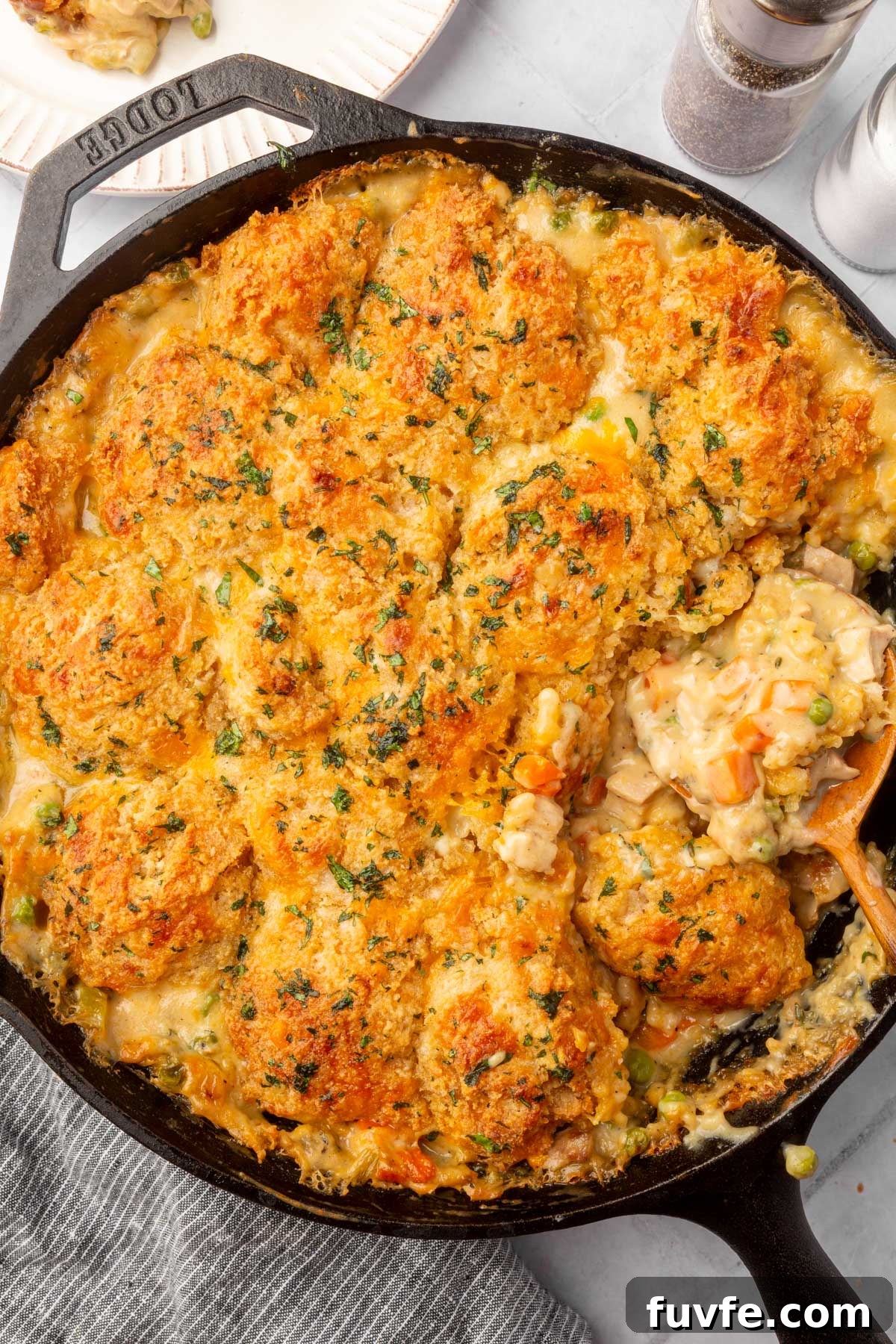 A cast iron skillet filled with golden-brown, biscuit-topped chicken pot pie, showing creamy filling with vegetables like carrots and peas, and a serving being scooped out with a wooden spoon.