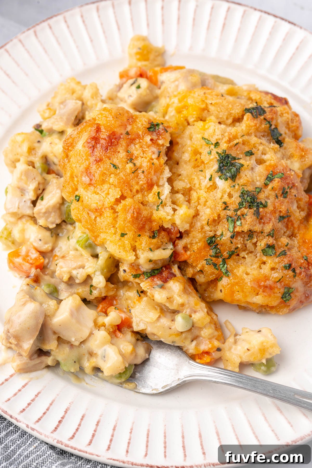 A plate with a serving of creamy chicken and vegetable casserole topped with golden, herb-baked biscuits. A fork rests on the plate, partially embedded in the casserole.