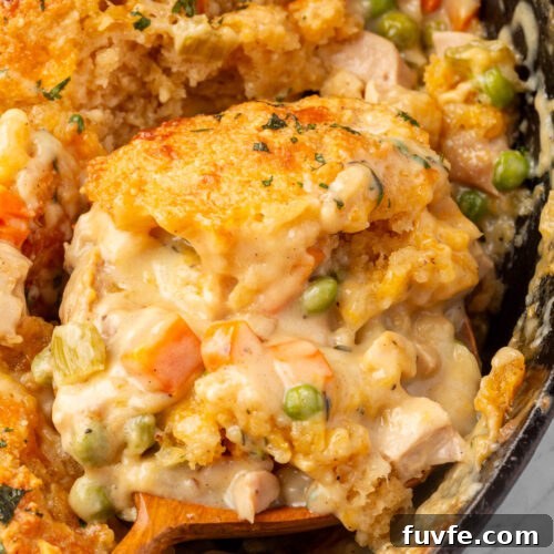 A close-up of a creamy chicken pot pie casserole with golden breadcrumb topping, peas, carrots, and chunks of chicken, being scooped with a wooden spoon.